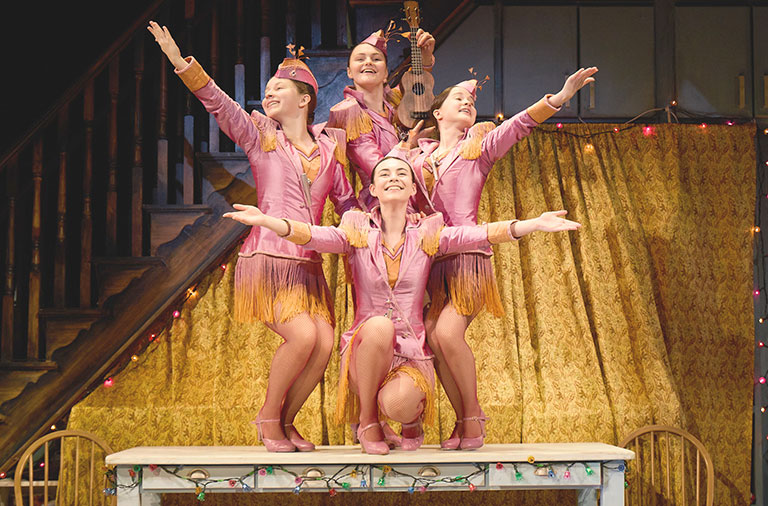 a photograph of 4 sisters, smiling brightly and dressed in matching outfits, performing as an old-time musical act, on top of a table in their family's home, which is decorated with Christmas lights