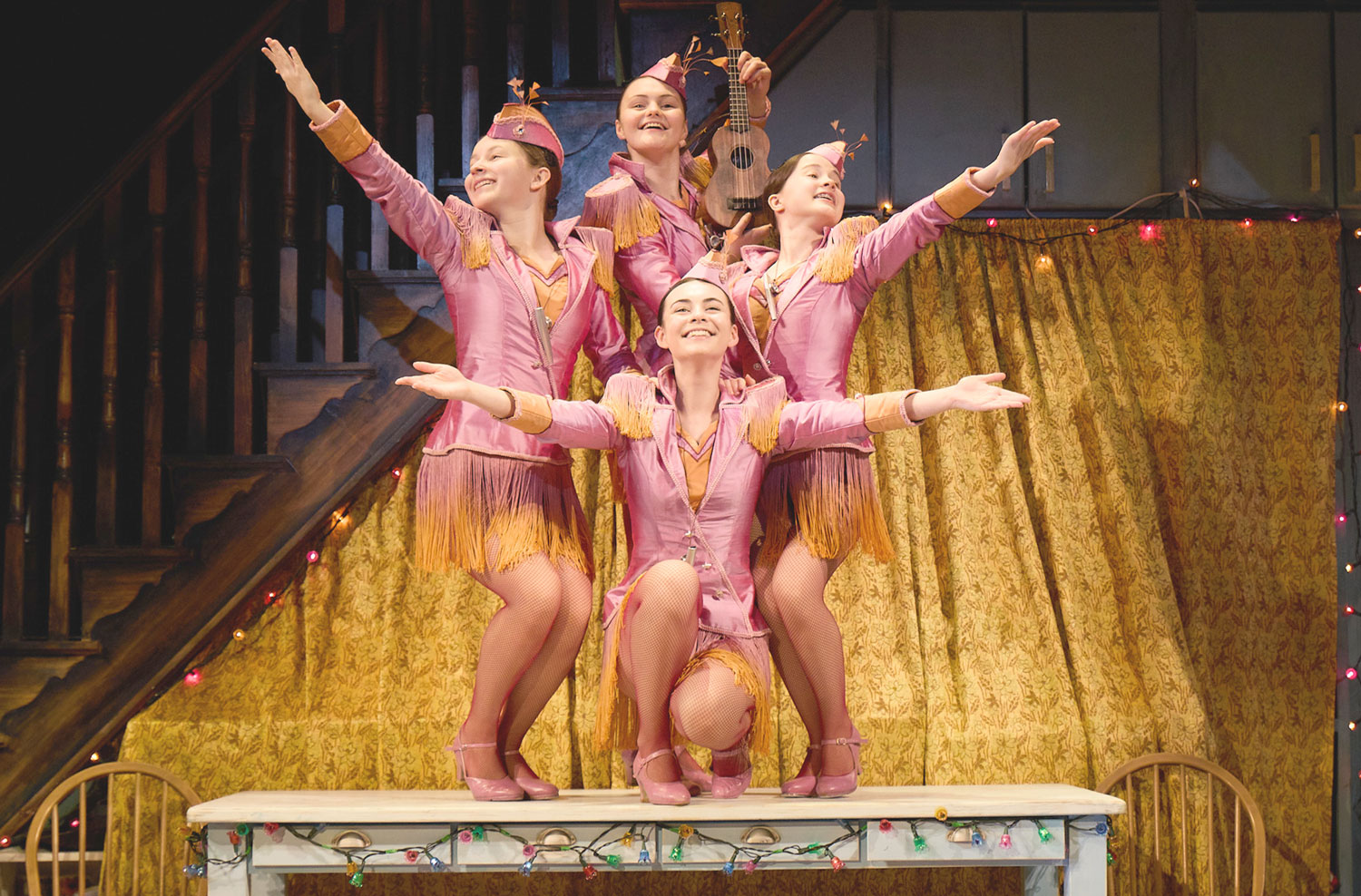 a photograph of 4 sisters, smiling brightly and dressed in matching outfits, performing as an old-time musical act, on top of a table in their family's home, which is decorated with Christmas lights